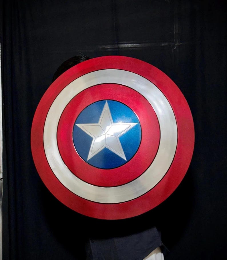 May include: A red, white, and blue shield with a silver star in the center. The shield has a circular design with concentric rings of red, white, and silver. The center is blue with a white star.