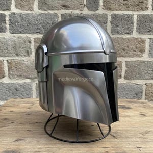 Op de afbeelding: Een zilverkleurige helm met een zwart vizier, op een zwarte draadstandaard. De helm heeft een kenmerkend hoekig ontwerp, met details zoals de wangkappen en de bovenkant van de helm. De tekst "medievalforgeco." is zichtbaar aan de zijkant.