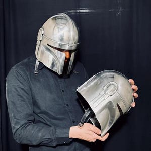 Może przedstawiać: Srebrny hełm Mandalorian z wizjerem w kształcie litery T i zaokrągloną g&oacute;rą. Hełm jest trzymany na tle ciemnego tła. Hełm ma sk&oacute;rzany pasek i jest wykonany z metalicznego materiału.