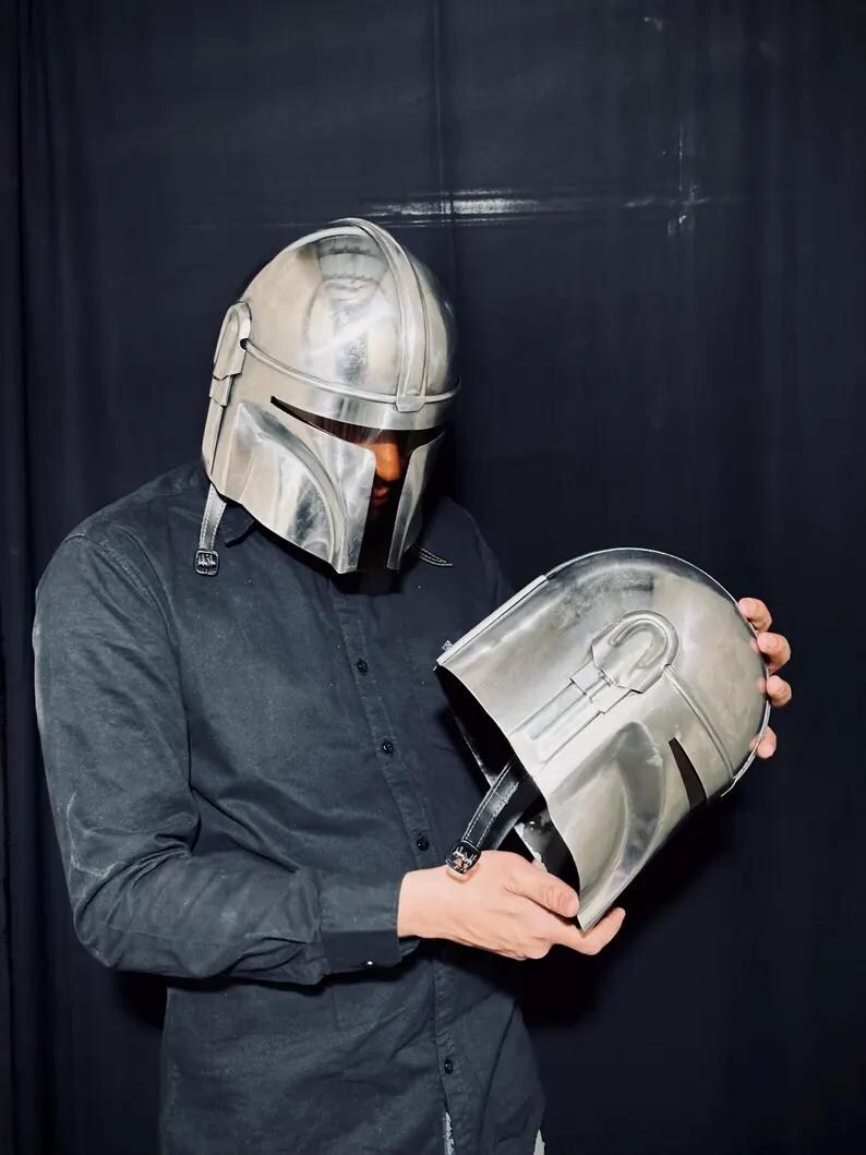 Może przedstawiać: Srebrny hełm Mandalorian z wizjerem w kształcie litery T i zaokrągloną g&oacute;rą. Hełm jest trzymany na tle ciemnego tła. Hełm ma sk&oacute;rzany pasek i jest wykonany z metalicznego materiału.