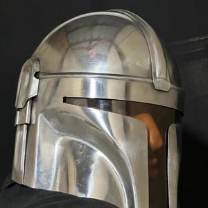 Może przedstawiać: Błyszczący, srebrny hełm o charakterystycznym designie, z zakrzywionym wizjerem i kątowymi detalami. Hełm wydaje się być wykonany z metalu i odbija otoczenie. Og&oacute;lna estetyka sugeruje motyw futurystyczny lub science fiction.