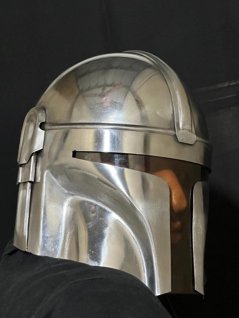 Może przedstawiać: Błyszczący, srebrny hełm o charakterystycznym designie, z zakrzywionym wizjerem i kątowymi detalami. Hełm wydaje się być wykonany z metalu i odbija otoczenie. Og&oacute;lna estetyka sugeruje motyw futurystyczny lub science fiction.