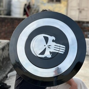 May include: A black and silver shield with a circular design. The center features a skull emblem. The shield has a silver border and a black center. The shield is a prop or collectible.