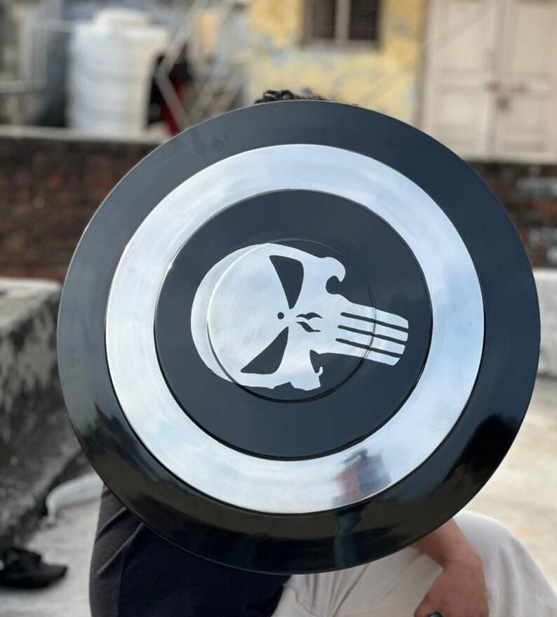 May include: A black and silver shield with a circular design. The center features a skull emblem. The shield has a silver border and a black center. The shield is a prop or collectible.