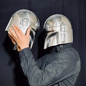 Może przedstawiać: Dwa błyszczące srebrne hełmy Mandalorian. Hełmy mają charakterystyczny wizjer w kształcie litery T i zaokrągloną g&oacute;rę z centralnym grzbietem. Prezentowane na czarnym tle.