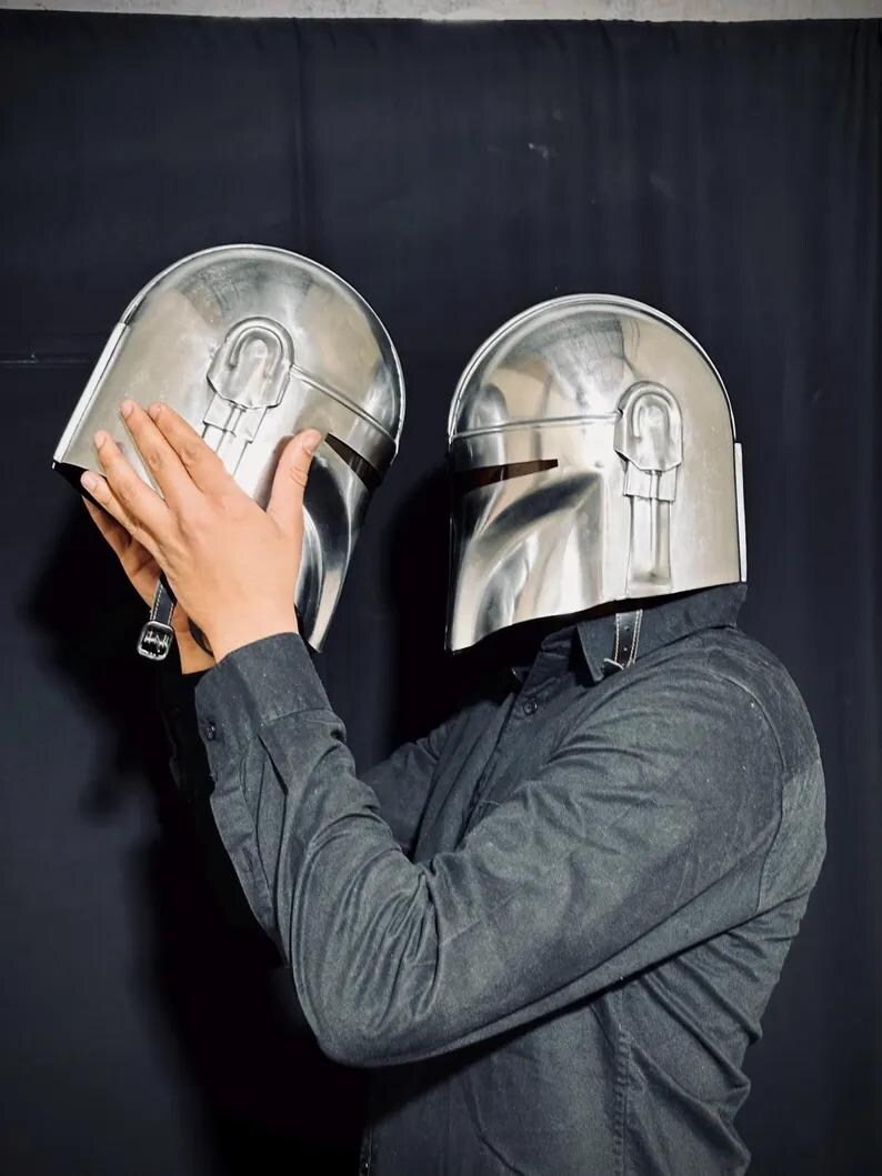 Może przedstawiać: Dwa błyszczące srebrne hełmy Mandalorian. Hełmy mają charakterystyczny wizjer w kształcie litery T i zaokrągloną g&oacute;rę z centralnym grzbietem. Prezentowane na czarnym tle.