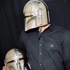 Może przedstawiać: Błyszczący, srebrny hełm Mandalorianina z wizjerem w kształcie litery T i zaokrągloną g&oacute;rą. Hełm trzyma osoba w czarnej koszuli. Drugi, mniejszy hełm trzymany jest w drugiej ręce osoby.