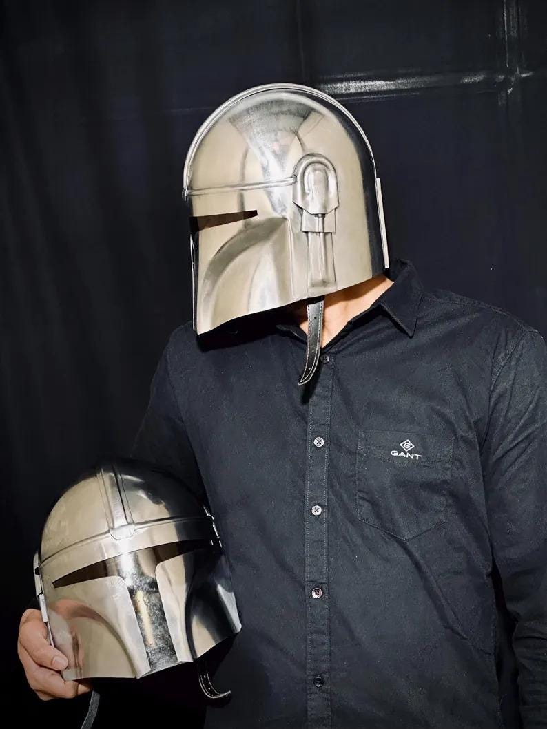 Może przedstawiać: Błyszczący, srebrny hełm Mandalorianina z wizjerem w kształcie litery T i zaokrągloną g&oacute;rą. Hełm trzyma osoba w czarnej koszuli. Drugi, mniejszy hełm trzymany jest w drugiej ręce osoby.