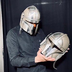 Może przedstawiać: Srebrny hełm Mandalorianina z pasującym drugim hełmem trzymanym w dłoniach. Hełmy mają metaliczne wykończenie i charakterystyczny wizjer w kształcie litery T. Tło to ciemna zasłona.