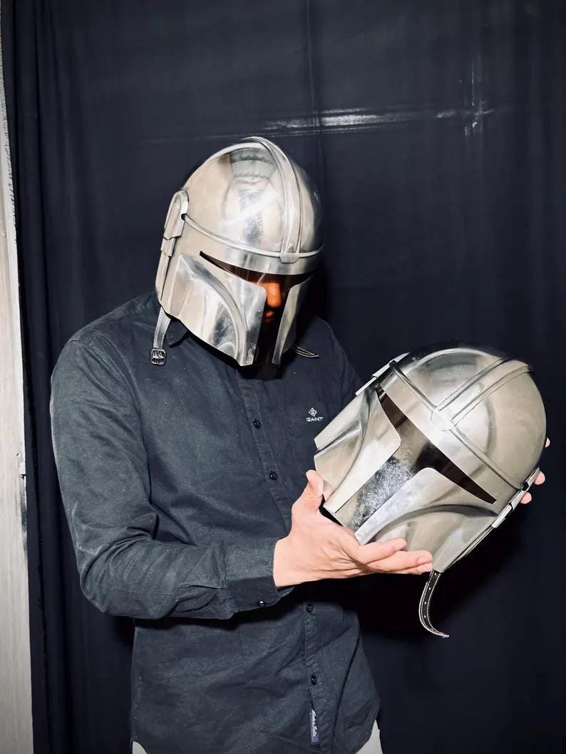 Może przedstawiać: Srebrny hełm Mandalorianina z pasującym drugim hełmem trzymanym w dłoniach. Hełmy mają metaliczne wykończenie i charakterystyczny wizjer w kształcie litery T. Tło to ciemna zasłona.