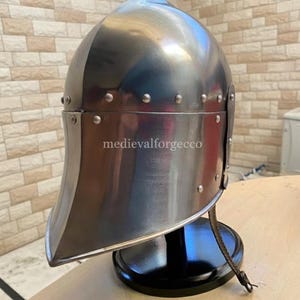 May include: A polished steel medieval helmet with a rounded top and a curved visor. The helmet is riveted and sits on a black stand. The text "medievalforgecco" is visible on the helmet.