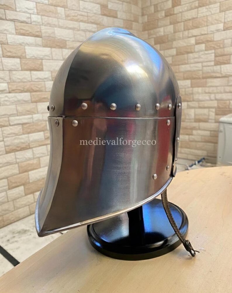 May include: A polished steel medieval helmet with a rounded top and a curved visor. The helmet is riveted and sits on a black stand. The text "medievalforgecco" is visible on the helmet.