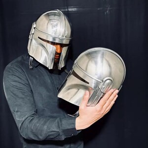 Może przedstawiać: Srebrny hełm Mandalorianina, z pasującym drugim hełmem trzymanym w dłoniach. Hełmy mają gładkie, metaliczne wykończenie i charakterystyczny kształt wizjera. Obraz jest na ciemnym tle.