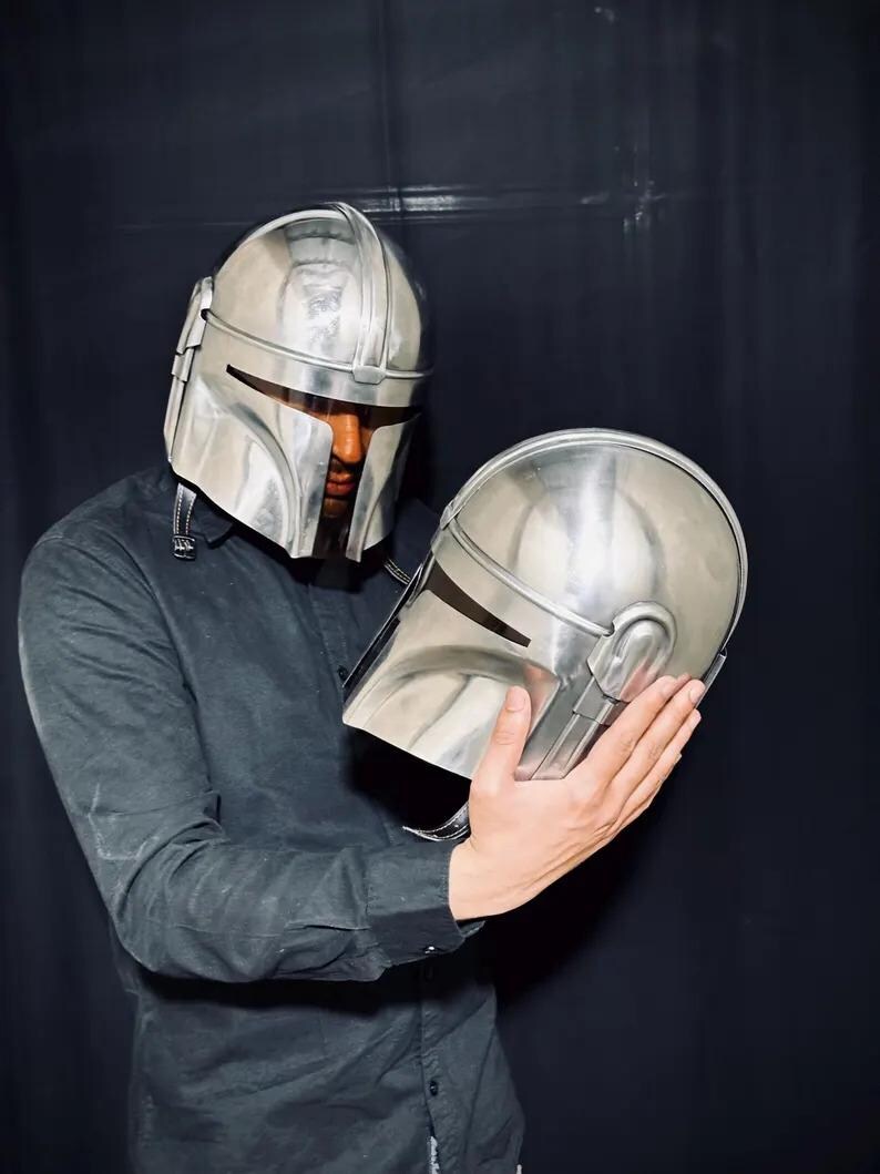 Może przedstawiać: Srebrny hełm Mandalorianina, z pasującym drugim hełmem trzymanym w dłoniach. Hełmy mają gładkie, metaliczne wykończenie i charakterystyczny kształt wizjera. Obraz jest na ciemnym tle.