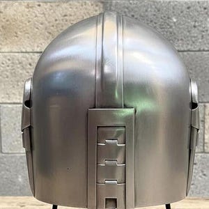Op de afbeelding: Een zilverkleurige helm met een afgeronde bovenkant en verticale ribbels. De achterkant van de helm heeft rechthoekige panelen met horizontale inkepingen. De helm wordt op een zwarte standaard gepresenteerd.
