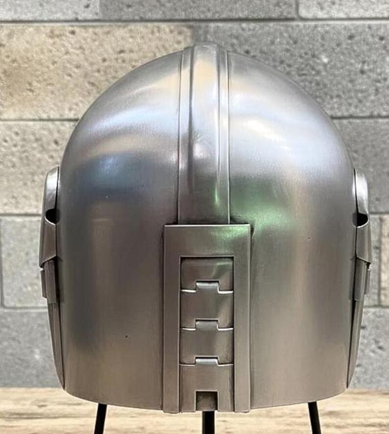 Op de afbeelding: Een zilverkleurige helm met een afgeronde bovenkant en verticale ribbels. De achterkant van de helm heeft rechthoekige panelen met horizontale inkepingen. De helm wordt op een zwarte standaard gepresenteerd.
