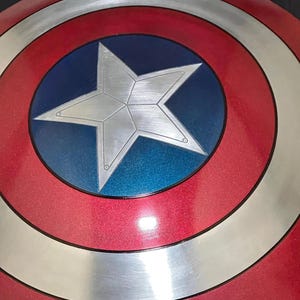 May include: A red, white, and blue shield with a large silver star in the center. The shield has concentric circles of red, silver, and red, with a blue circle in the middle. A classic superhero accessory.