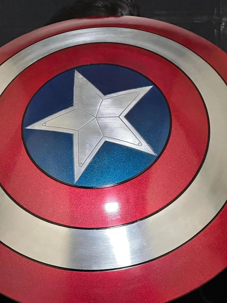 May include: A red, white, and blue shield with a large silver star in the center. The shield has concentric circles of red, silver, and red, with a blue circle in the middle. A classic superhero accessory.