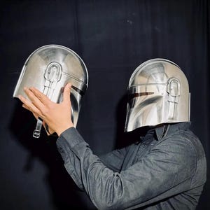 Może przedstawiać: Dwa srebrne hełmy Mandalorian. Jeden hełm jest trzymany przed drugim. Hełmy mają wizjer i zaokrągloną g&oacute;rę z pionowym detalem paska. Osoba na zdjęciu ma na sobie ciemnoszarą koszulę zapinaną na guziki.
