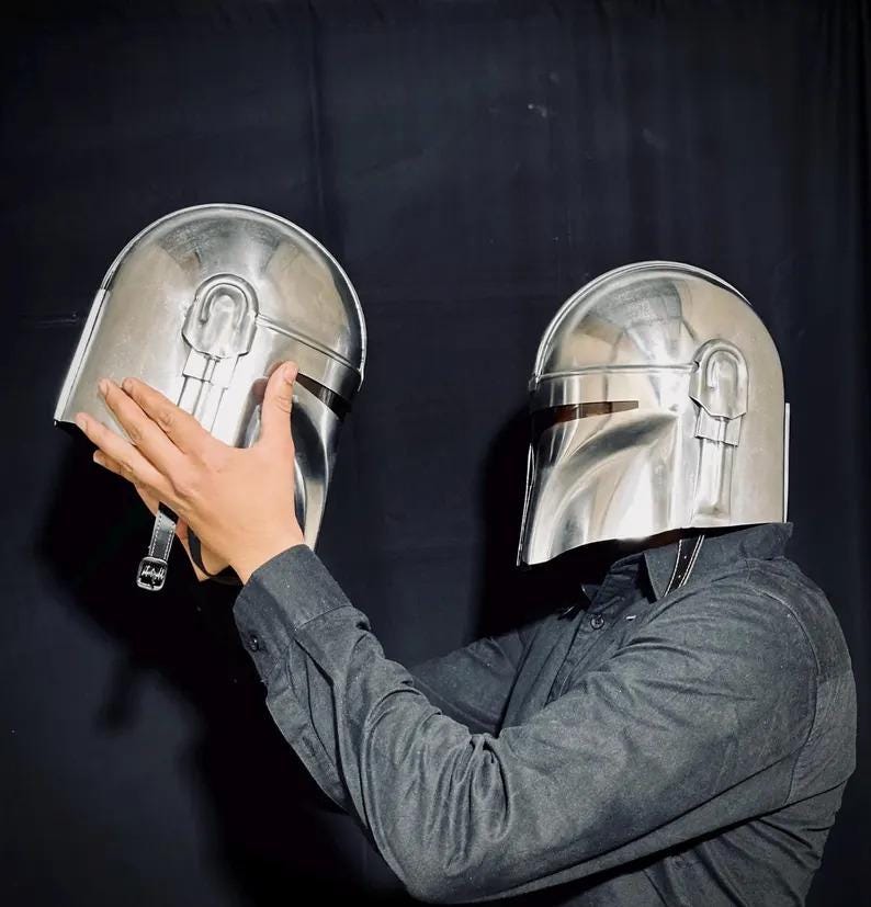 Może przedstawiać: Dwa srebrne hełmy Mandalorian. Jeden hełm jest trzymany przed drugim. Hełmy mają wizjer i zaokrągloną g&oacute;rę z pionowym detalem paska. Osoba na zdjęciu ma na sobie ciemnoszarą koszulę zapinaną na guziki.