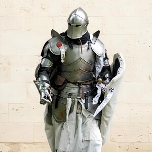 May include: A complete set of medieval armour, featuring a helmet, breastplate, and gauntlets. The silver-coloured armour is detailed with rivets and layered plates. A white cloth is draped over the armour, and a shield is visible. The background is a light-coloured stone wall.