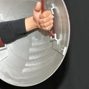 May include: A silver and red shield with a red handle is held by a person. The shield has concentric circles and metal brackets. The handle is made of a red strap. The person is wearing a black long-sleeve shirt.