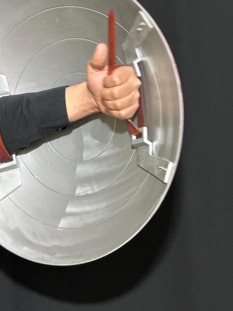May include: A silver and red shield with a red handle is held by a person. The shield has concentric circles and metal brackets. The handle is made of a red strap. The person is wearing a black long-sleeve shirt.