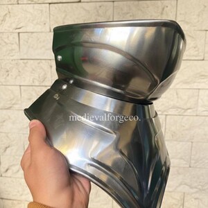 May include: A polished, silver-colored metal gorget, a piece of medieval armor, held by a person. The gorget has a curved design to protect the neck and upper chest. The text "medievalforgeco." is visible on the armor.