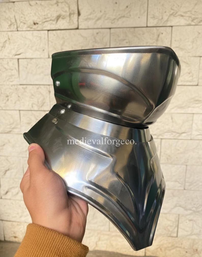 May include: A polished, silver-colored metal gorget, a piece of medieval armor, held by a person. The gorget has a curved design to protect the neck and upper chest. The text "medievalforgeco." is visible on the armor.