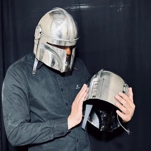 Może przedstawiać: Srebrny hełm Mandalorian z wizjerem w kształcie litery T i pasującym hełmem trzymanym w dłoniach. Hełmy mają metaliczne wykończenie i są prawdopodobnie wykonane z metalu lub podobnego materiału. Tło jest ciemne.