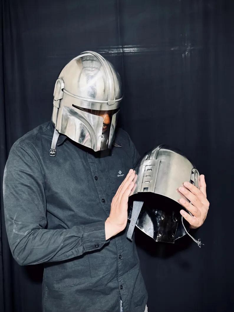 Może przedstawiać: Srebrny hełm Mandalorian z wizjerem w kształcie litery T i pasującym hełmem trzymanym w dłoniach. Hełmy mają metaliczne wykończenie i są prawdopodobnie wykonane z metalu lub podobnego materiału. Tło jest ciemne.