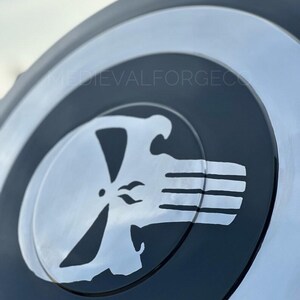 May include: A black and silver shield featuring a white skull emblem. The shield has a circular design with concentric rings of black and silver. The skull emblem is in the center, with a stylized skull design.