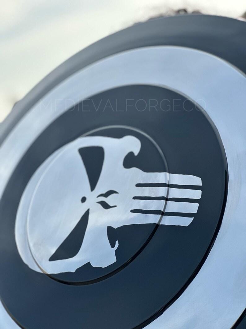 May include: A black and silver shield featuring a white skull emblem. The shield has a circular design with concentric rings of black and silver. The skull emblem is in the center, with a stylized skull design.