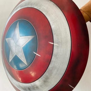 May include: A red, white, and blue shield with a silver star in the center. The shield has a circular design with concentric rings of color. The shield is a prop or collectible item.