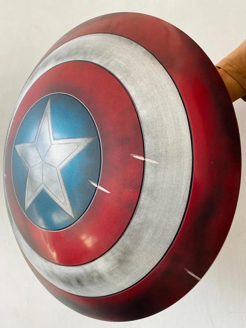 May include: A red, white, and blue shield with a silver star in the center. The shield has a circular design with concentric rings of color. The shield is a prop or collectible item.