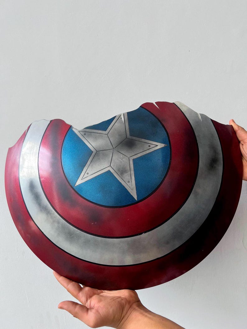 May include: A Captain America shield with a distressed finish. The shield features a blue center with a white star, surrounded by red and silver concentric circles. The shield has a damaged edge.