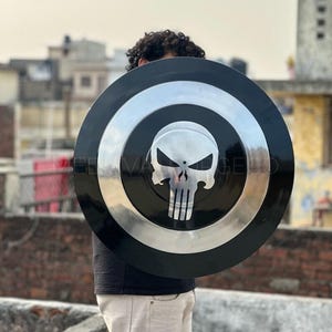 May include: A black and silver shield with a skull emblem. The shield has a circular design with a black outer ring, a silver inner ring, and a skull graphic in the center. The shield is held by a person.