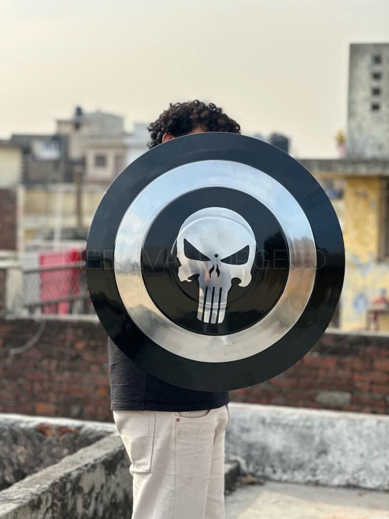 May include: A black and silver shield with a skull emblem. The shield has a circular design with a black outer ring, a silver inner ring, and a skull graphic in the center. The shield is held by a person.