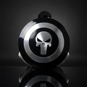 May include: A black and silver shield featuring a skull emblem. The shield has a circular design with a black center and silver outer rings. The skull emblem is in the center of the shield.