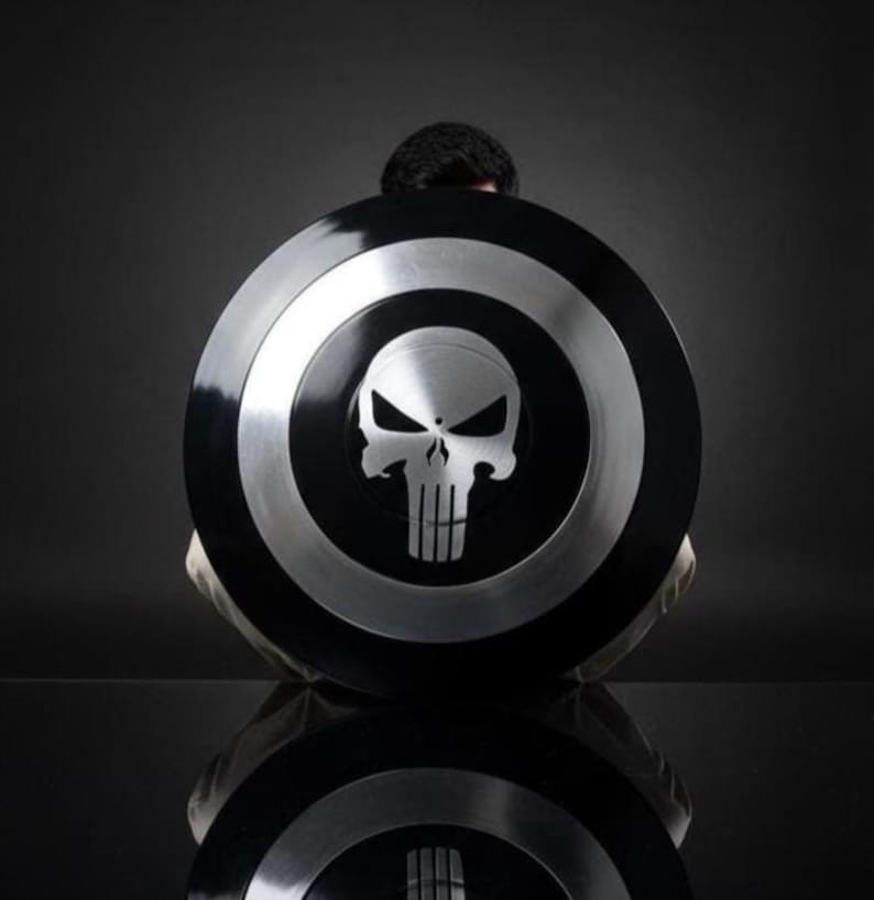 May include: A black and silver shield featuring a skull emblem. The shield has a circular design with a black center and silver outer rings. The skull emblem is in the center of the shield.