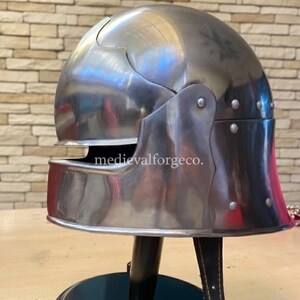May include: A polished, silver-colored medieval helmet with a rounded top and a visor. The helmet has a hinged visor and cheek plates, and is mounted on a black stand. The text "medievalforgeco." is visible on the helmet.
