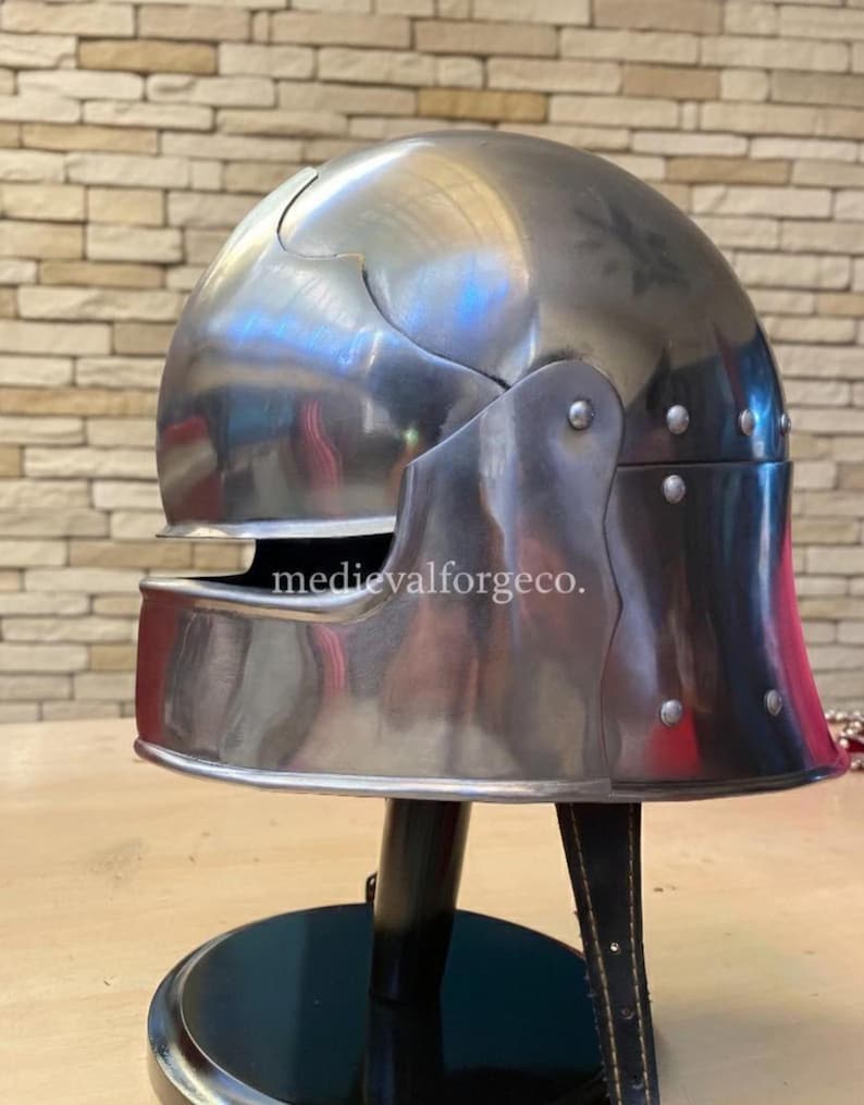 May include: A polished, silver-colored medieval helmet with a rounded top and a visor. The helmet has a hinged visor and cheek plates, and is mounted on a black stand. The text "medievalforgeco." is visible on the helmet.