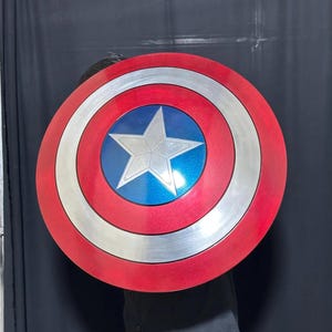 May include: A round shield with a red, white, and blue color scheme. The shield features a silver star in the center, surrounded by a blue circle, white ring, and red outer ring. The shield is a prop or collectible.