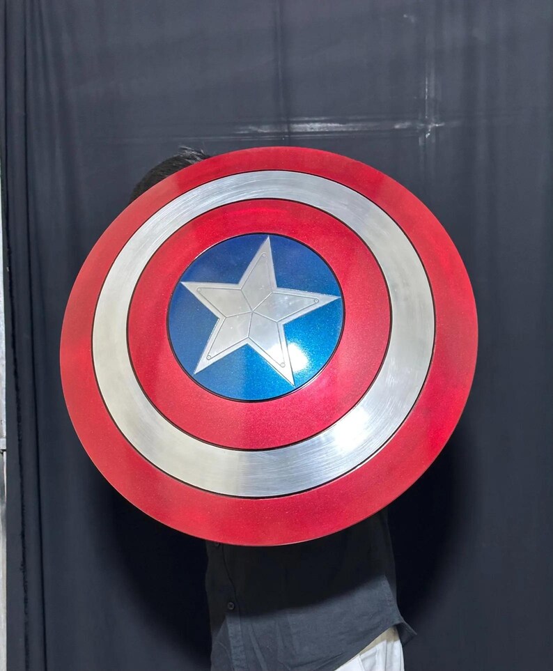 May include: A round shield with a red, white, and blue color scheme. The shield features a silver star in the center, surrounded by a blue circle, white ring, and red outer ring. The shield is a prop or collectible.