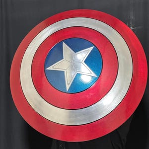 May include: A round shield with a red, silver, and blue color scheme. The shield features a silver star in the center, surrounded by concentric rings of silver, white, and red. The shield is approximately 24 inches in diameter.
