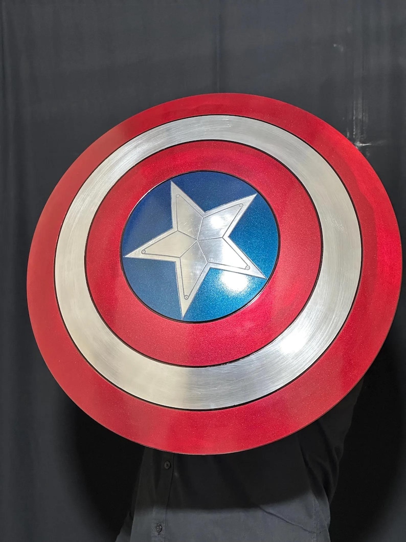 May include: A round shield with a red, silver, and blue color scheme. The shield features a silver star in the center, surrounded by concentric rings of silver, white, and red. The shield is approximately 24 inches in diameter.
