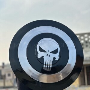 May include: A black and silver shield featuring a white skull emblem. The shield has a circular design with a black outer ring, a silver inner ring, and a black center with the skull logo. The text "MEDIEVAL FORGE CO" is visible.