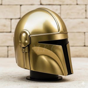 Op de afbeelding: Een goudkleurige Mandalorian helm, gepresenteerd op een zwarte standaard. De helm heeft een gladde, metallic afwerking met een T-vormig vizier en gedetailleerde zijkanten. De helm is een replica van de iconische helm uit de Star Wars franchise.