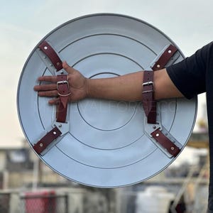 May include: A light gray, circular shield with concentric rings and brown leather straps. The shield has a metallic appearance and is designed with riveted straps for secure handling. The shield is a prop or decorative item.
