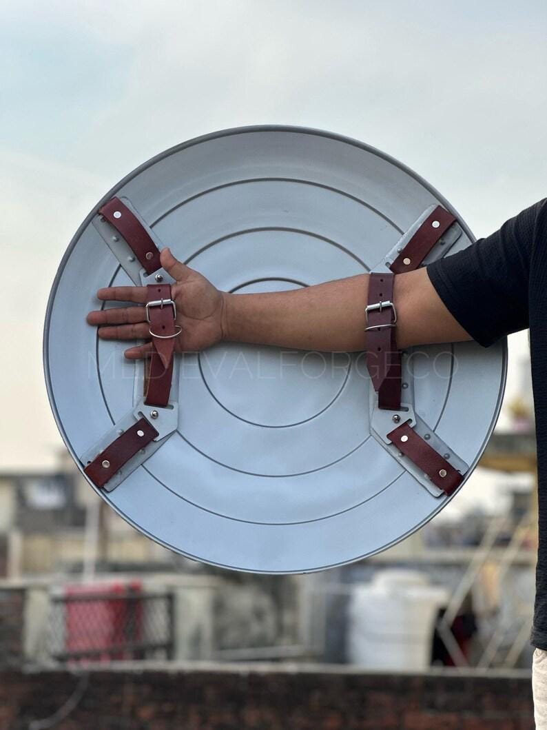 May include: A light gray, circular shield with concentric rings and brown leather straps. The shield has a metallic appearance and is designed with riveted straps for secure handling. The shield is a prop or decorative item.